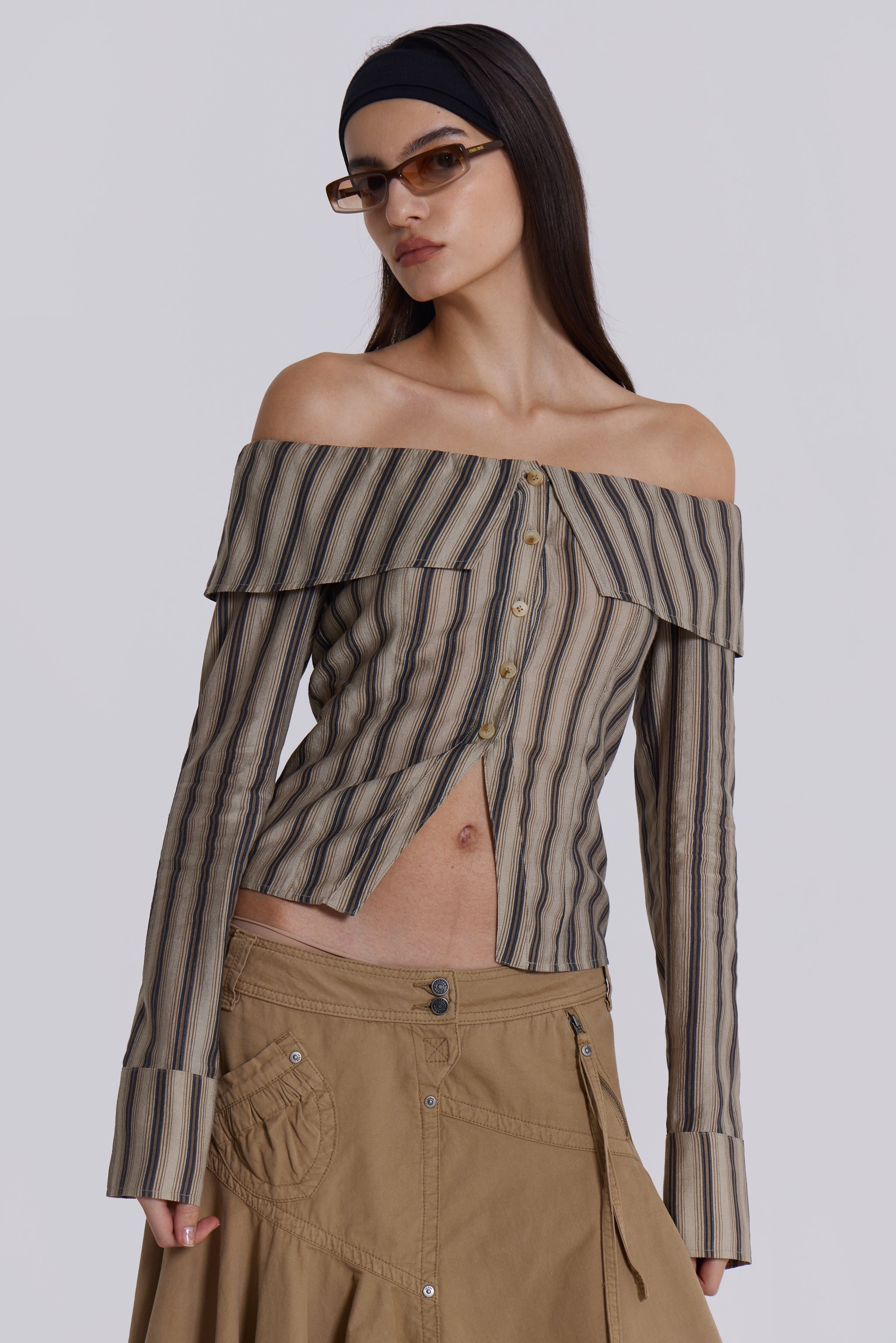  Female model wearing a khaki and brown striped off the shoulder long sleeve shirt with flared cuffs. Styled with a beige choppy cargo skirt. 