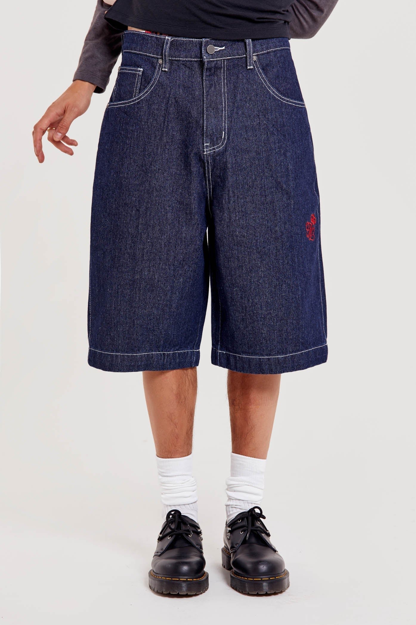Male wearing indigo blue denim jorts in a jumbo fit with JL embroidered logo and Union Jack start branding on back pocket. 