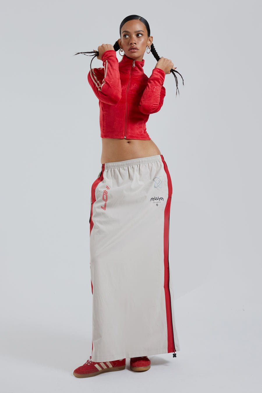 Female wearing red logo monogram embossed high neck long sleeve velour track top. Styled with almond windbreaker maxi length skirt.